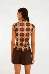 Top de manga corta con cuello camisero y cierre frontal de botones, confeccionado en tejido liviano con estampado geométrico de flores en tonos marrón, naranja y beige.