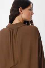 Camisa de corte oversize color chocolate, con cuello clásico, cierre frontal de botones y mangas largas con puños abotonados. Presenta un diseño fluido y holgado con sutiles frunces en los hombros.