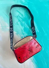 Bandolera Kaira de la línea Ottenta de Carolina De Cunto, confeccionada en cuero color rojo con diseño estilo splash en negro y bolsillo frontal superior en tono cobre. Cuenta con correa larga regulable negra con el nombre de la marca en blanco y cierre superior con cremallera.