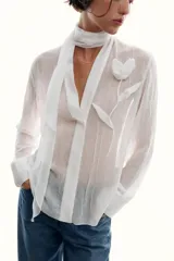 Blusa de tejido semitransparente con efecto arrugado, escote en pico y manga larga. Presenta un detalle decorativo frontal con estructura en forma de flor.