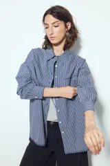 Camisa de silueta amplia y caída fluida, color celeste con rayas verticales blancas. Presenta cuello camisero clásico, cierre frontal con botones y un bolsillo aplicado en el pecho. Las costuras están resaltadas en azul oscuro.