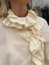 Camisa de manga larga color marfil, con volados en el cuello, en la parte frontal y en los puños, además de mangas abullonadas y puños elásticos fruncidos.