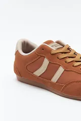 Zapatilla urbana de perfil bajo con diseño retro, confeccionada en poliuretano con detalles en textura tipo gamuza. Presenta puntera redondeada, cordones clásicos y suela de goma color caramelo.