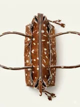 Cartera de tamaño medio confeccionada en cuero vacuno con textura de pelo y estampado animal print tipo vaca. Presenta estructura rígida, doble asa de hombro, cierre superior, detalle de tira horizontal con tachas y hebillas metálicas, y lazos decorativos en los laterales.