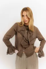 Camisa de estilo romántico confeccionada en algodón, con cuello alto, volados frontales y mangas largas con puños acampanados también decorados con volados.