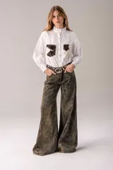 Camisa blanca de manga larga con cuello alto gorguera con volados y detalles de alforzas verticales en el frente. Cuenta con dos bolsillos en el pecho de pelo con estampado de vaca en tonos marrones y blancos.