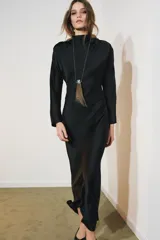 Vestido largo negro de tejido satinado, con cuello subido, manga larga y drapeado en los hombros. Presenta una abertura en la espalda con cierre de botones forrados a tono.