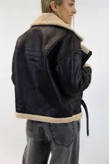 Campera oversize de efecto cuero color marrón con interior, solapas, cuello y puños de corderito sintético en tono tostado. Cuenta con detalles de tiras ajustables con hebilla en el cuello, mangas y ruedo, y cierre frontal cruzado.