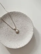 Collar de cadena fina tipo serpiente en tono plateado con un dije circular que presenta una perla central rodeada por pequeñas piedras brillantes.