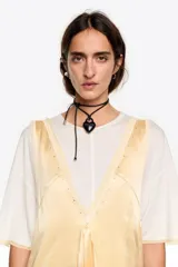 Vestido midi de diseño bimateria con sobrevestido de satén color amarillo claro. Presenta escote en V profundo con lazo frontal, detalles de encaje en el ruedo y aberturas laterales con cordones. Se combina con una base de remera blanca de manga corta y cuello redondo.