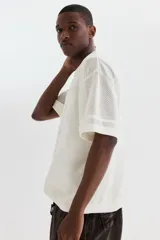 Remera de malla color blanco hueso, de manga corta, con cuello redondo y hombros caídos. Presenta un diseño de tejido perforado con un número grande grabado en el frente.