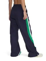 Pantalón deportivo de corte holgado y pierna ancha, confeccionado en tejido de nailon con textura arrugada. Presenta un diseño en color azul marino con paneles laterales en verde y blanco, cintura elástica y botones a presión en los laterales de las piernas.