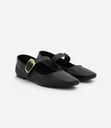 Zapatilla tipo bailarina de cuero negro, con punta redondeada, tacón bajo y una tira decorativa en el empeine con hebilla metálica dorada.