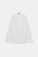 Camisa de popelín de algodón a rayas verticales, con cuello subido y aplicación de tejido tipo pañuelo anudado al frente. Presenta manga larga con puños anchos y cierre frontal con botones ocultos.