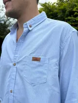 Camisa de manga larga a rayas celestes y blancas, con cuello abotonado, bolsillo en el pecho con etiqueta de cuero sintético y botones de madera.