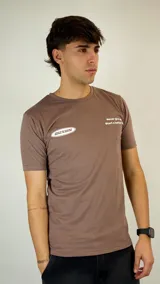 Remera de algodón color marrón con cuello redondo y mangas cortas. Presenta un estampado gráfico en el pecho con el logo de la marca en un óvalo y una frase motivacional.