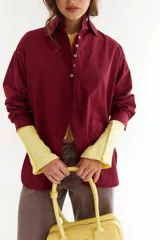 Camisa de algodón color bordó, con cuello clásico, cierre frontal de botones y diseño con recorte diagonal en el frente y aberturas laterales. Presenta un bordado en zigzag en la espalda.