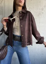 Chaqueta de gamuza color chocolate, con corte recto, cuello clásico y cierre frontal con botones. Presenta un diseño calado decorativo en el ruedo y bolsillos frontales tipo parche.