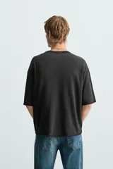 Camiseta de punto con corte boxy fit, cuello redondo y manga corta. Color negro con efecto lavado y terminaciones irregulares. Presenta un estampado frontal con texto y un gráfico circular en tonos blanco y rojo.