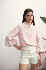 Camisa a rayas verticales color beige y blanco, con cuello clásico, cierre frontal con botón dorado, mangas largas abullonadas con puños elásticos, dos bolsillos frontales y corte holgado.