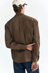 Camisa marrón de manga larga, confeccionada en sarga suave con cuello inglés, cierre frontal de botones, bolsillo en el pecho y bajo redondeado.