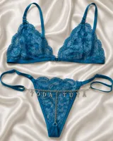 Conjunto de lencería azul compuesto por un bralette de encaje con argolla central y una tanga a juego adornada con una cadena metálica con dijes de letras que forman una frase y correas regulables.