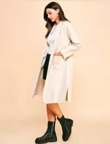 Cardigan largo de tejido color beige claro, sin cuello, con bolsillos frontales y detalles de pespuntes decorativos a lo largo del borde frontal.