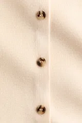 Saco corto de punto suave en color beige, con diseño de hombros caídos y escote amplio ribeteado. Cuenta con mangas largas y cierre frontal de botones con efecto carey.