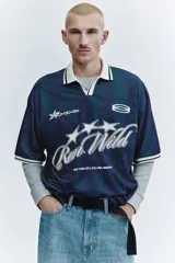 Camiseta de fútbol de manga corta en malla estampada, con diseño a rayas verticales en tonos azul y verde oscuro. Cuenta con cuello polo blanco y escote en V con ribete negro, además de estampados gráficos frontales con estrellas y la inscripción "Run Wild". Presenta un corte holgado.
