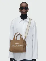Bolso tote Marc Jacobs modelo The Small Tote de lona marrón tostado, con asas superiores y correa ajustable de hombro color crema. Presenta el texto estampado "THE TOTE BAG MARC JACOBS" en color blanco en la parte frontal.