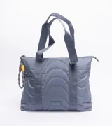 Cartera tipo tote bag de diseño acolchado con costuras curvas, color azul grisáceo. Cuenta con doble asa de hombro y un detalle de cordón decorativo con terminales en amarillo y gris en el lateral.
