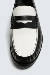 Mocasín de cuero liso, estilo penny loafer, con contraste de color blanco y negro. Presenta un antifaz superior con una abertura central y costuras visibles en el borde. Tiene suela negra de bajo tacón.