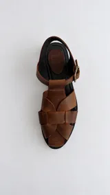 Sandalia de cuero marrón con diseño trenzado artesanal en el empeine, pulsera ajustable al tobillo con hebilla metálica y suela plana.