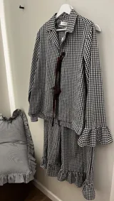 Conjunto de dos piezas de pijama confeccionado en algodón a cuadros vichy rosados y blancos. Incluye una camisa de manga larga con puños acampanados y cierre delantero con cordones para atar, y un pantalón largo de corte recto con volados en el ruedo.