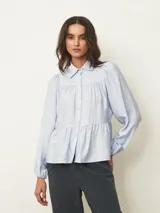 Camisa de corte holgado y diseño escalonado, color celeste, con cuello camisero, cierre frontal de botones y mangas largas con puños elásticos.