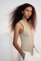 Top de algodón color beige con escote en V, sin mangas y cierre frontal con botones. Presenta un diseño asimétrico en el bajo con abertura delantera.