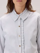 Camisa de algodón color gris claro, con cuello clásico y detalle de volados en la cartera frontal. Presenta mangas largas con puños abotonados y un corte recto.
