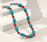 Collar de cuentas asimétrico compuesto por piezas alargadas de imitación piedra turquesa con vetas negras, intercaladas con pequeñas cuentas esféricas de color rojo. Incluye cadena extensora metálica en tono dorado.