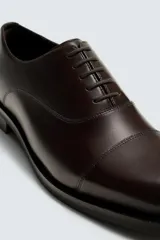Zapato de vestir tipo oxford con puntera reforzada, confeccionado en piel lisa de color negro. Presenta cierre de cordones, forro interior y plantilla de piel, y suela con tacón bajo.