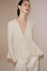 Blusa de viscosa color rosado con cuello redondo, manga larga abullonada con puños elásticos y cierre frontal con lazadas. Presenta un diseño con volantes en el bajo y corte asimétrico.