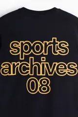 Remera boxy de algodón color negro, con cuello redondo acanalado, mangas cortas y hombros caídos. Presenta un pequeño estampado amarillo en el pecho con el texto "sports archives 08".