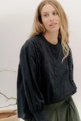 Camisa de algodón blanca con cuello a la base, cierre frontal con botones y mangas largas con puños delicados. Presenta volados verticales y detalles de cintas de terciopelo en contraste a lo largo del frente.