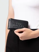 Billetera Marc Jacobs The Top Zip Multi Wallet de cuero negro granulado, con cierre superior dorado y la inscripción "THE WALLET MARC JACOBS" grabada en relieve en el frente.