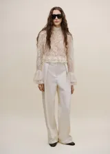 Blusa de encaje color crema con cuello alto, mangas largas con volados en los puños y detalle de volado en el ruedo.