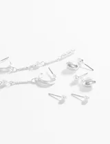 Set de caravanas plateadas que incluye: aros tipo argolla con cadena larga y perlas colgantes, aros tipo ear cuff con cadena y perla colgante, aros colgantes con forma de corazón liso y perla, y dos pares de aros tipo stud con perla pequeña.