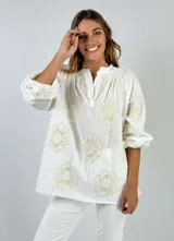 Blusa blanca de corte holgado con mangas tres cuartos abullonadas, cuello mao con pequeña abertura frontal y bordados florales en hilo dorado.