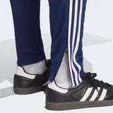 Pantalón deportivo Adidas Tiro 25 Essentials, color azul marino, con corte clásico y puños ajustados. Presenta las tres franjas características de Adidas en blanco a lo largo de las costuras laterales y el logo de la marca en blanco en el muslo.