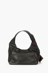 Cartera de cuero vacuno color negro, con costuras a contratono en color blanco y un práctico bolsillo frontal desmontable en color bordó con tiras envolventes. Cuenta con asa para el hombro.