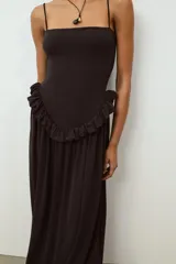 Vestido largo de color negro con escote recto y tirantes finos ajustables. Presenta un diseño con detalle de volante tipo peplum en la cintura y falda con caída fluida.