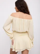 Vestido corto color crema, de diseño off-shoulder con mangas largas abullonadas, cintura ajustada con frunces elásticos y falda corta de volados.